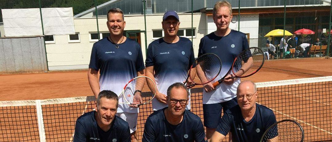 Eine Gruppe von fünf Männern posiert für ein Foto auf einem Tennisplatz, jeder hält Tennisschläger in der Hand. Sie tragen aufeinander abgestimmte blaue Hemden und Shorts. Hinter ihnen befindet sich ein Netz und ein Gebäude mit Regenschirmen.
