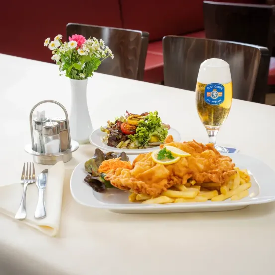 Ein Tisch für eine Mahlzeit mit einem Teller mit gebratenem Fisch, Pommes Frites, einem Salat, einem Glas Bier und einer Vase mit Blumen.