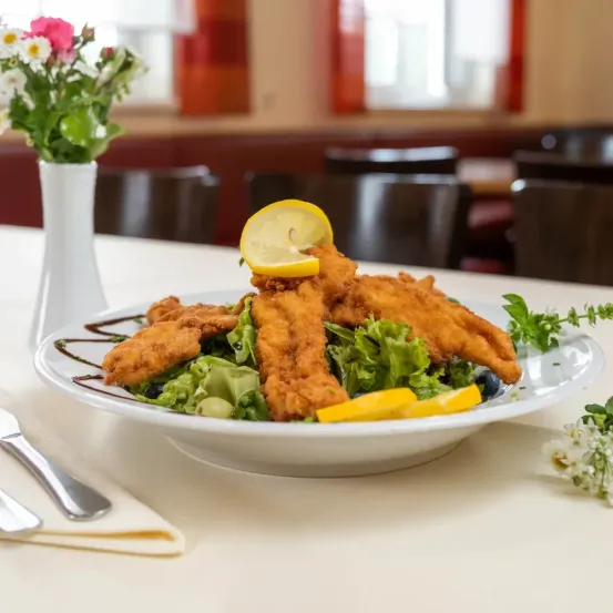 Ein Teller mit gebratenen Fischfilets, Zitronenscheiben und Salat, begleitet von einer Blumenvase auf einer weißen Tischdecke.