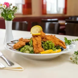 Ein Teller mit gebratenen Fischfilets, Zitronenscheiben und Salat, begleitet von einer Blumenvase auf einer weißen Tischdecke.