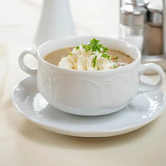 Eine weiße Schüssel mit cremiger Suppe, garniert mit Schlagsahne und grünen Kräutern, auf einem weißen Unterteller auf einem Tisch.