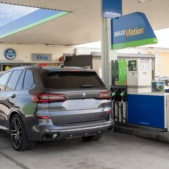 Ein grauer BMW SUV ist an einer Tankstelle mit einem MAXX Motion-Schild geparkt. Das Heck des Autos ist sichtbar und zeigt seine Rücklichter und Auspuffrohre.