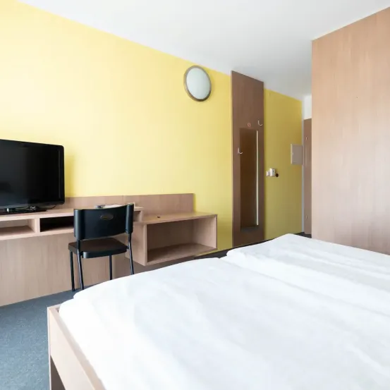 Ein modernes Hotelzimmer mit einem Bett, einem Schreibtisch, einem Stuhl und einem Flachbildfernseher an der Wand. Das Zimmer hat eine gelbe Wand und eine Holztür.