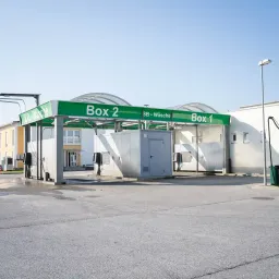 Eine öffentliche Waschanlage mit zwei Boxen, die mit Box 1 und Box 2 beschriftet sind, unter einem grünen Dach, auf dem SB-Wäsche steht.
