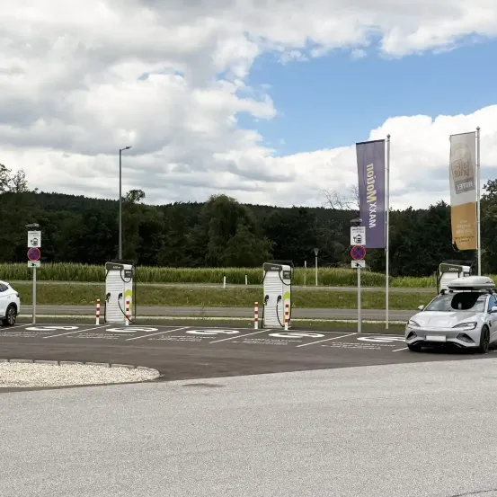 Ein Parkplatz mit Elektroauto-Ladestationen, einem weißen Auto mit Dachgepäckträger und einem Banner mit dem Text 'Mobilität XXL'.