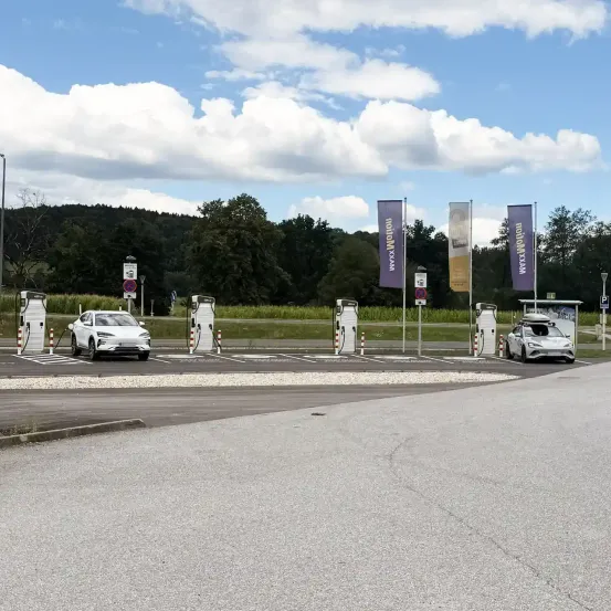Ein Parkplatz im Freien mit Elektroauto-Ladestationen, zwei Autos laden, und ein klarer blauer Himmel mit Wolken.
