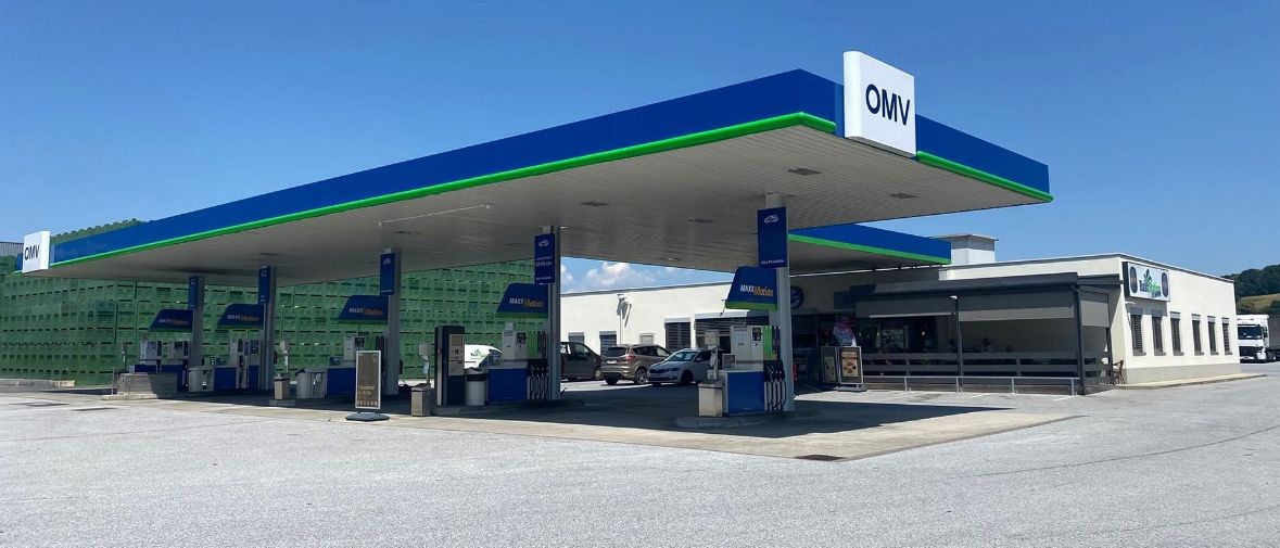 Eine OMV-Tankstelle mit blauer und weißer Beschilderung, mehrere geparkte Autos und ein Gebäude mit einer Bank und sitzenden Personen.