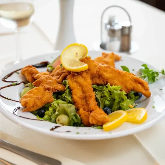 Ein Teller mit frittierten Hähnchenstreifen, grünem Salat, überbacken mit Zitronenscheiben und mit Sauce beträufelt, begleitet von einem Weinglas und Gewürzen.