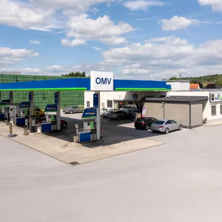 Eine Tankstelle mit blauer und weißer Beschilderung, die OMV beschriftet ist, mit mehreren geparkten Autos und einem Gebäude im Hintergrund.