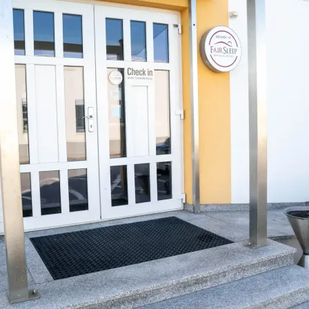 Der Eingang zu einem Hotel mit einem Schild, das 'Check in' und ein rundes Schild mit dem Text 'Fair Sleep' auf einer gelben Wand zeigt.
