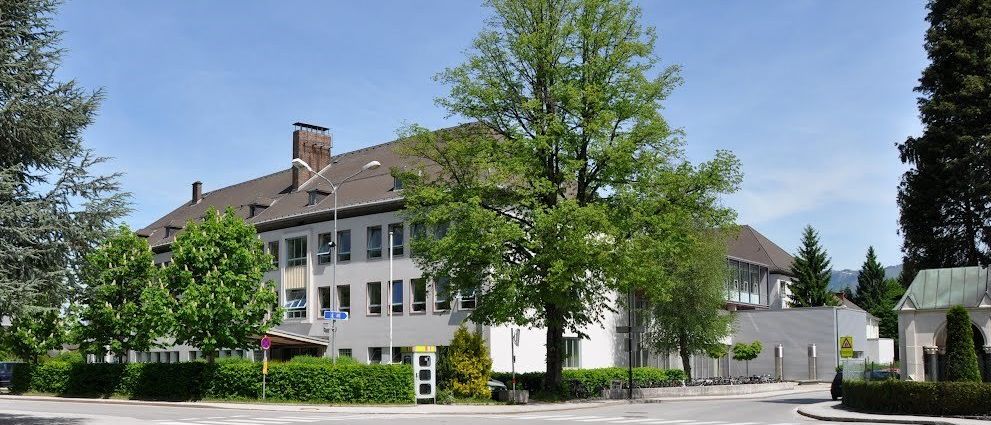 Ein großes Gebäude mit vielen Fenstern, einem Schornstein und einem Baum davor, unter einem klaren blauen Himmel.