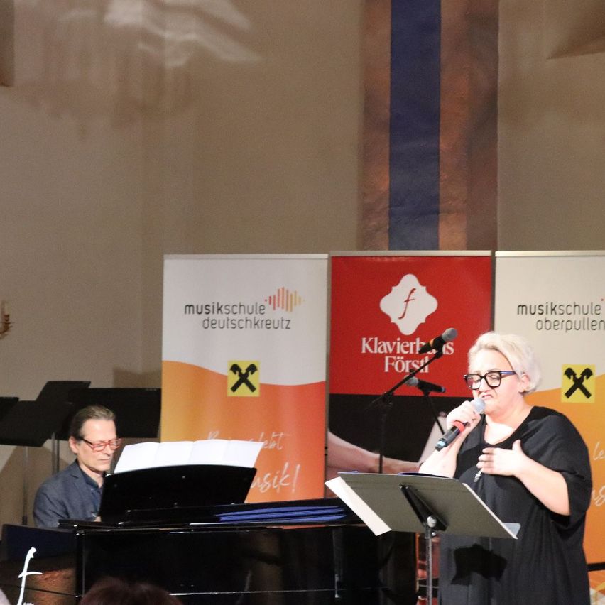 Eine Frau singt in ein Mikrofon, während sie vor einem Klavier steht. Ein Mann spielt das Klavier hinter ihr. Sie sind beide bei einer Veranstaltung der Musikschule.