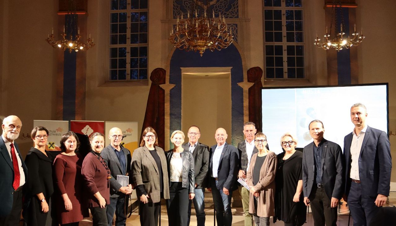 Eine Gruppe von formell gekleideten Menschen steht vor einer Bühne mit einer Projektionsleinwand. Eine Kronleuchter hängt von der Decke.