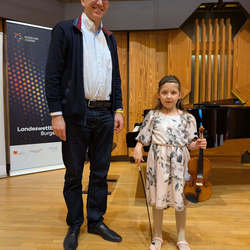 A man and a young girl stand near a piano in a music room, the girl holds a violin and looks at the camera, while the man smiles at her.