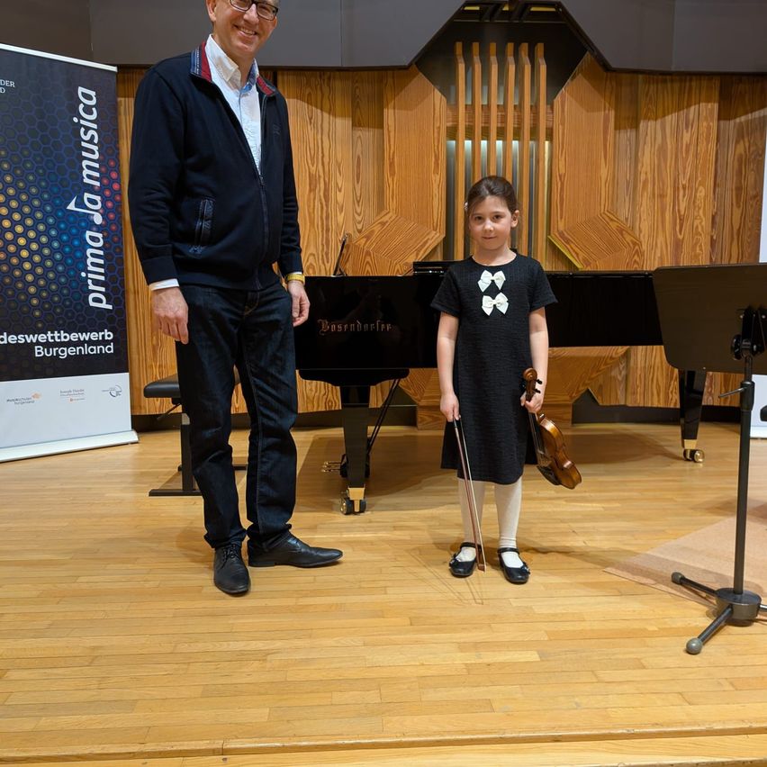 A man and a young girl stand on a stage in front of a piano. The man smiles and the girl holds a violin.