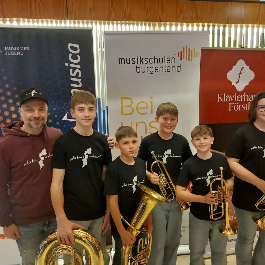 Six young boys and two adults stand in front of a backdrop with musical instruments, smiling and holding brass instruments. They wear matching black t-shirts. Behind them, three banners are displayed, one featuring a logo and text.