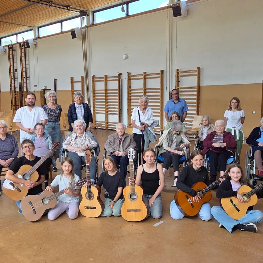 Eine Gruppe von Menschen, einige im Rollstuhl, posiert für ein Foto mit Gitarren. Einige sitzen auf dem Boden, während andere hinter ihnen stehen. Der Raum hat Holzböden, weiße Wände und Holzleitern an der Wand.