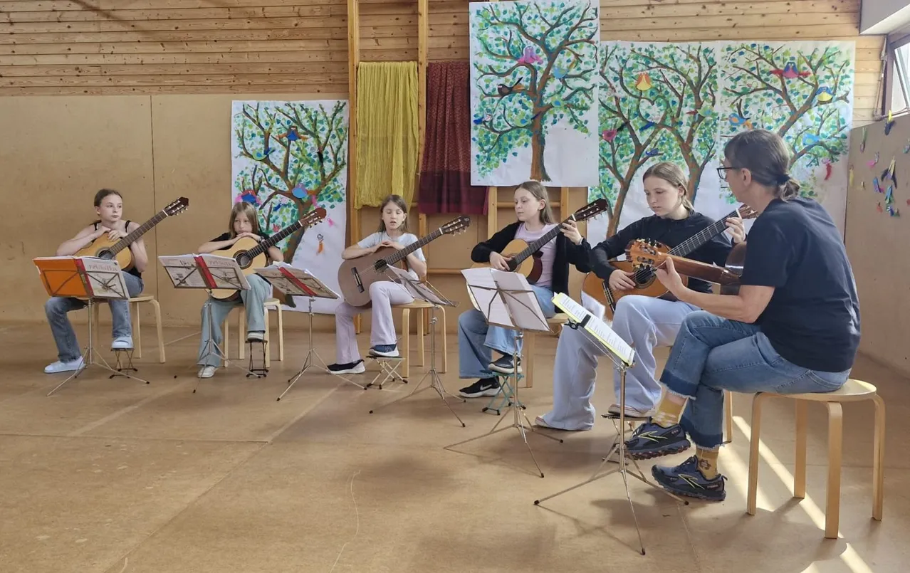 Eine Gruppe junger Mädchen in einem Musikraum übt das Gitarrespielen, mit Notenständern und einer hölzernen Wand mit Gemälden im Hintergrund.