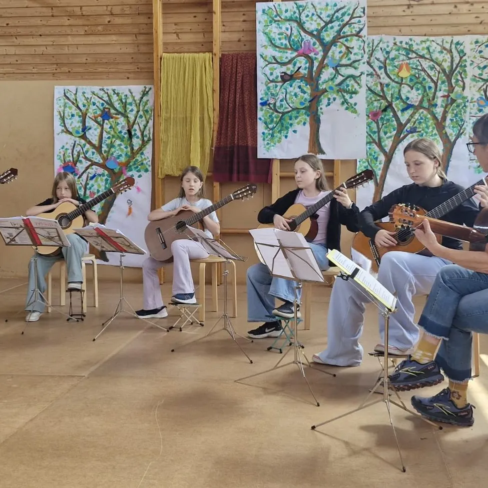 Eine Gruppe junger Mädchen in einem Musikraum übt das Gitarrespielen, mit Notenständern und einer hölzernen Wand mit Gemälden im Hintergrund.