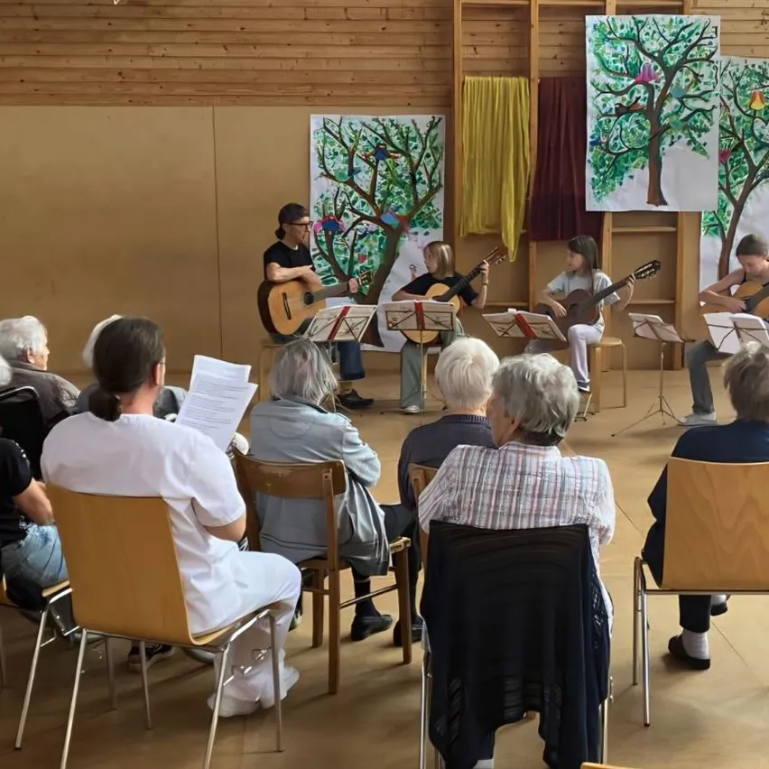 Eine Gruppe von Menschen sitzt auf Stühlen und beobachtet eine Darbietung von vier Gitarristen, von denen einer auf einem Hocker sitzt. Die Künstler haben Notenblätter vor sich.