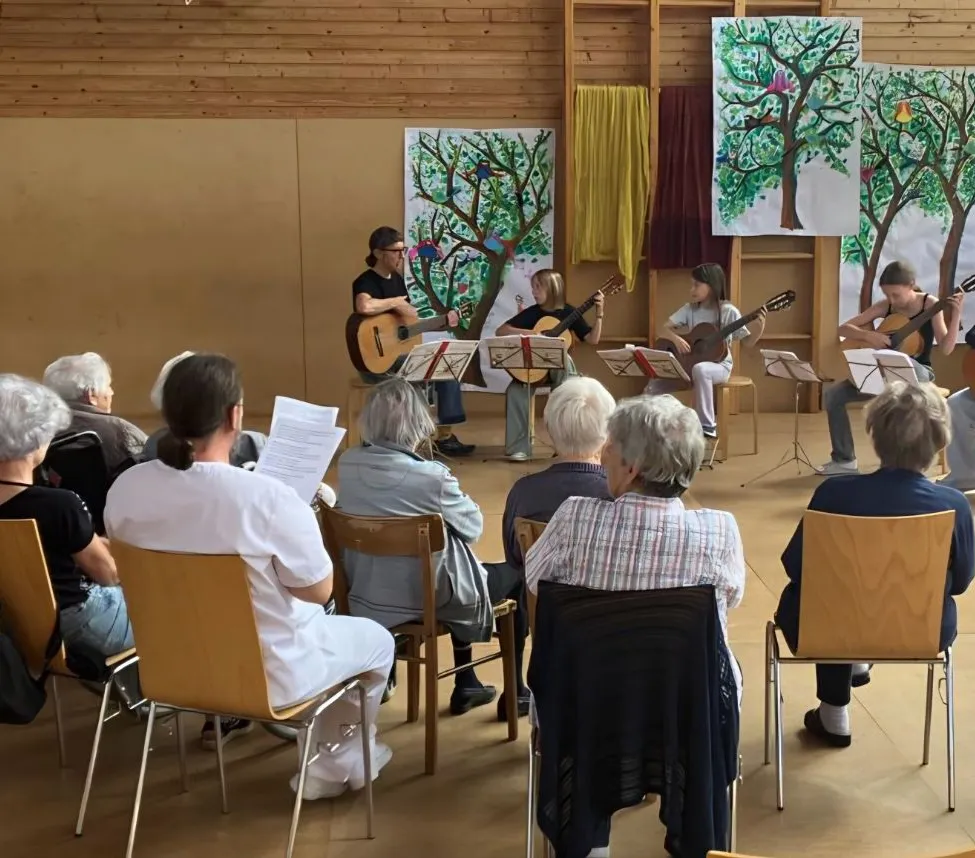 Eine Gruppe von Menschen sitzt auf Stühlen und beobachtet eine Darbietung von vier Gitarristen, von denen einer auf einem Hocker sitzt. Die Künstler haben Notenblätter vor sich.