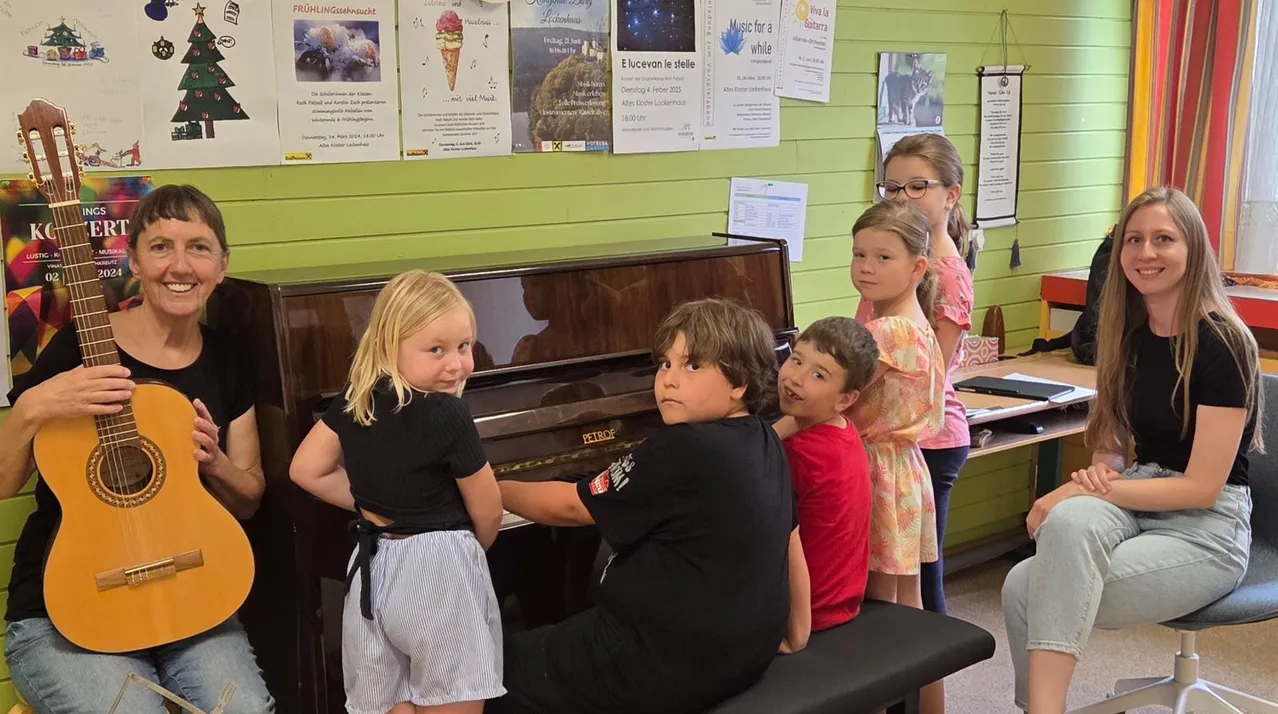 Vier Kinder sitzen auf einer Klavierbank, wobei zwei Mädchen daneben stehen. Ein Mädchen lächelt, während es Klavier spielt. Hinter ihnen befinden sich Poster an einer grünen Wand.