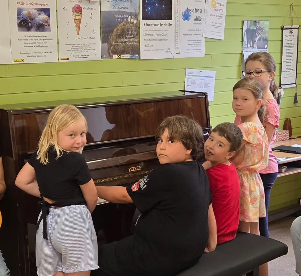 Vier Kinder sitzen auf einer Klavierbank, wobei zwei Mädchen daneben stehen. Ein Mädchen lächelt, während es Klavier spielt. Hinter ihnen befinden sich Poster an einer grünen Wand.