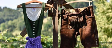 Traditional Bavarian clothing hangs on a wooden pole in a vineyard. The image features a green vest and brown lederhosen with a purple skirt. The text reads 'Fest am Ploschitzberg 29. August 2026 Beginn 17:00 Uhr.'