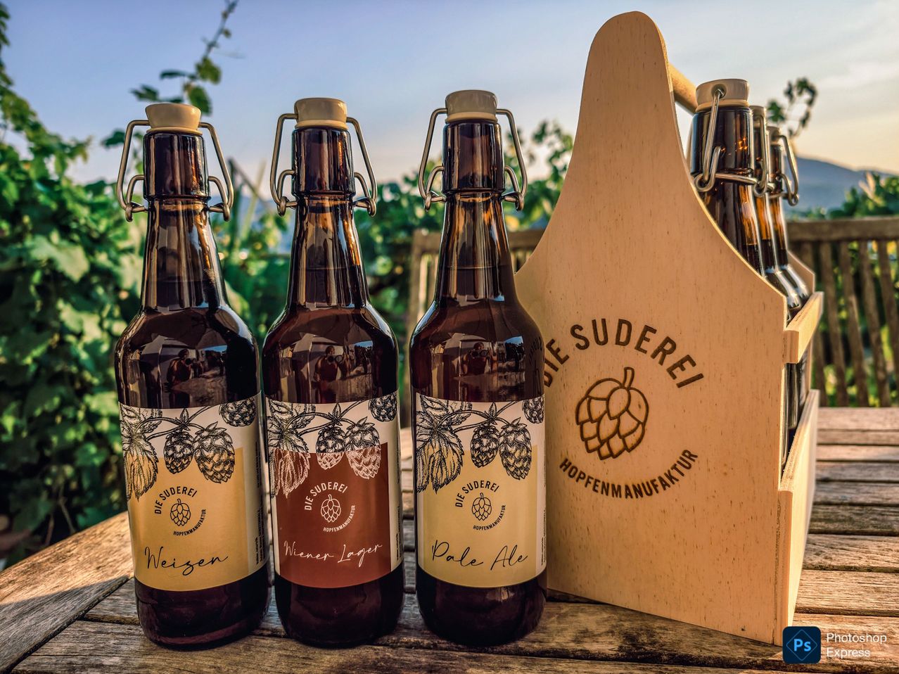Three bottles of beer with brown labels and metal caps are displayed on a wooden table. The labels read 'Weizen', 'Wiener Lager', and 'Pale Ale'. A wooden box with the text 'Die Suderei' is also present.