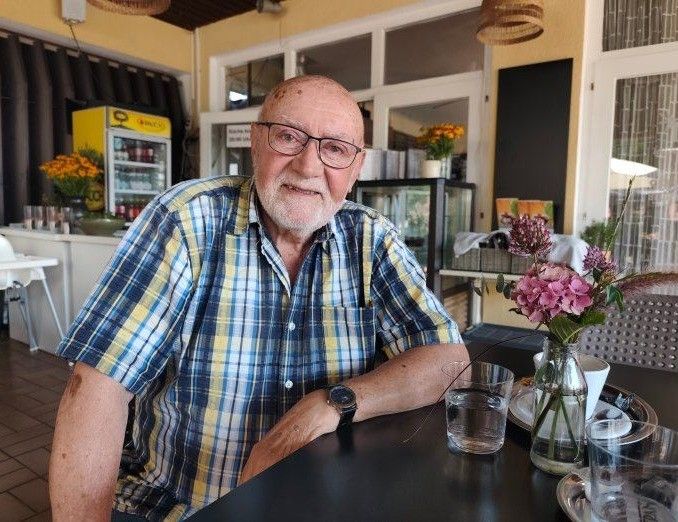An elderly man with glasses and a plaid shirt is sitting at a table in a restaurant, smiling and looking at the camera. There is a vase of flowers, glasses, and a plate on the table. Behind him, there is a refrigerator, a shelf with items, and a blackboard.