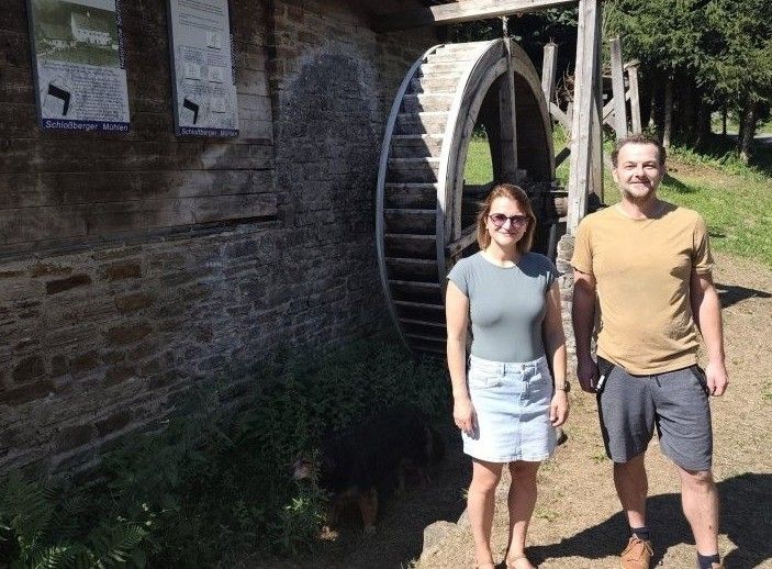 A man and a woman stand next to an old water wheel, smiling for the camera. A dog is sitting on the ground nearby.