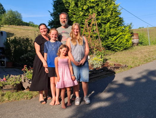 A family of five stands on a road, smiling for a photo. They are surrounded by greenery, including trees and plants. The background features a grassy field and a distant hill.