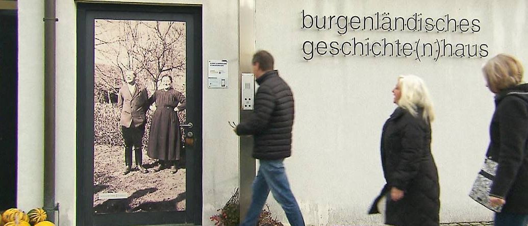 Zwei Menschen gehen an einem Gebäude vorbei. Ein Foto eines Mannes und einer Frau steht an der Tür. An der Wand steht 'Burgenländische Geschichte (n)h.'