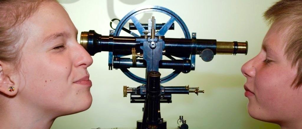 Zwei Frauen schauen durch ein altes Teleskop mit grünem Hintergrund. Die Frau auf der linken Seite hat die Augen geschlossen, während die Frau auf der rechten Seite den Mund offen hat.