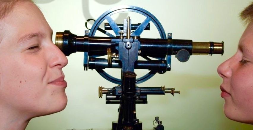 Zwei Frauen schauen durch ein altes Teleskop mit grünem Hintergrund. Die Frau auf der linken Seite hat die Augen geschlossen, während die Frau auf der rechten Seite den Mund offen hat.