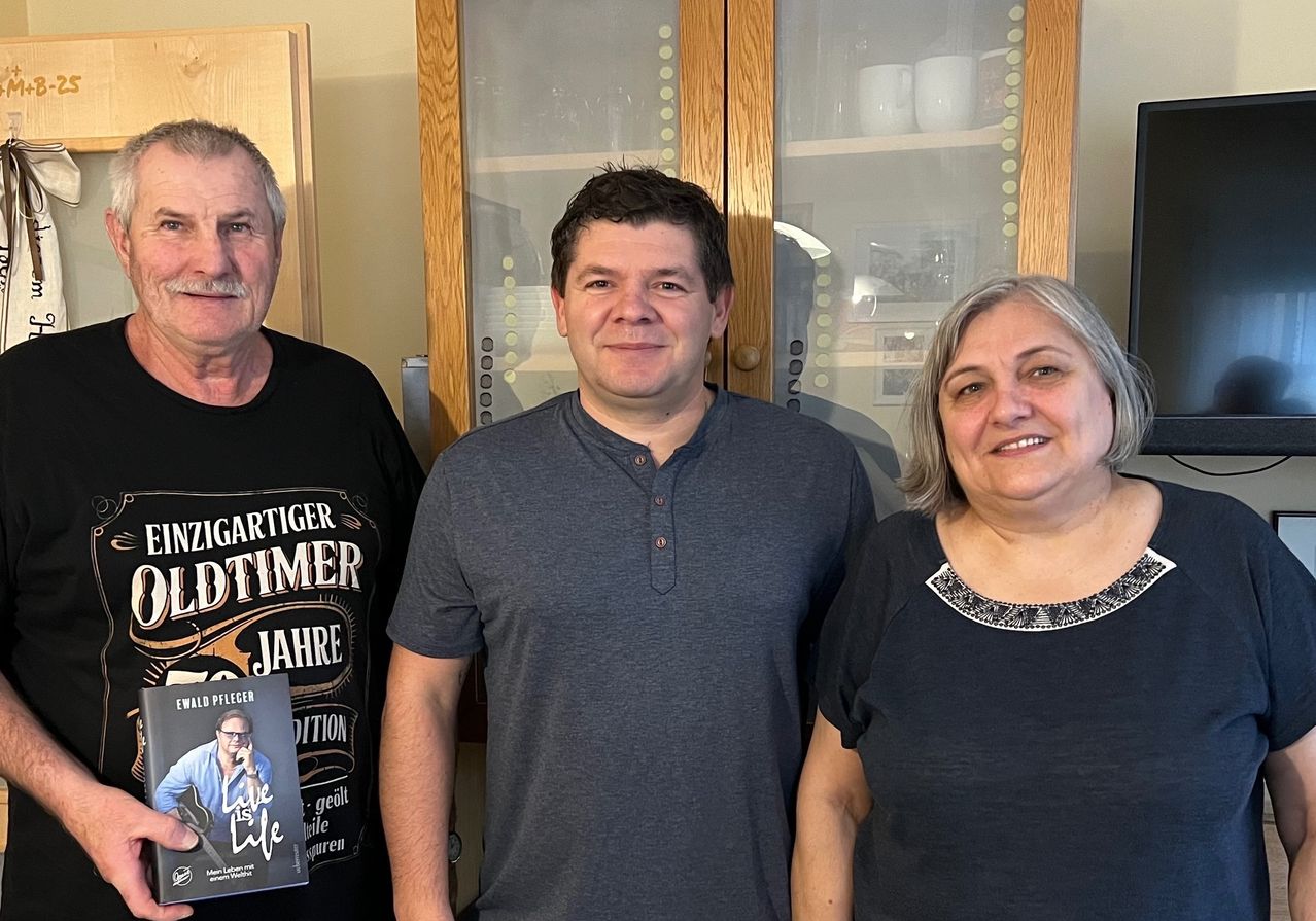 Drei Personen stehen in einem Raum mit einem Schrank, der Mann auf der linken Seite hält ein Buch, der Mann in der Mitte trägt ein graues Hemd und die Frau auf der rechten Seite trägt ein schwarzes Oberteil.