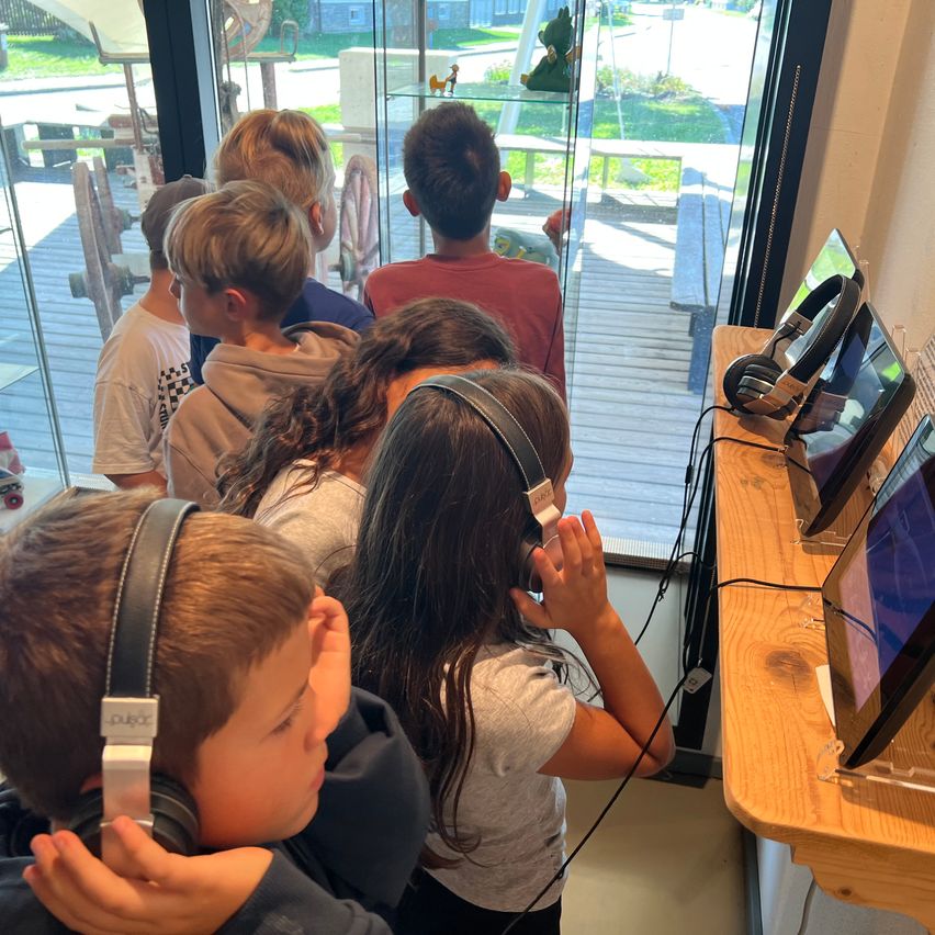 Eine Gruppe von Kindern mit Kopfhörern versammelt sich um einen Monitor, möglicherweise in einem Klassenzimmer oder Lernumgebung.
