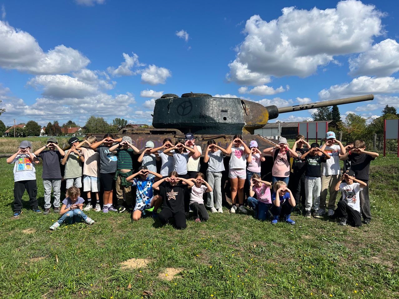 Eine Gruppe von Kindern und Erwachsenen posiert für ein Foto vor einem alten Panzer auf einem Grasfeld unter einem teilweise bewölkten Himmel.