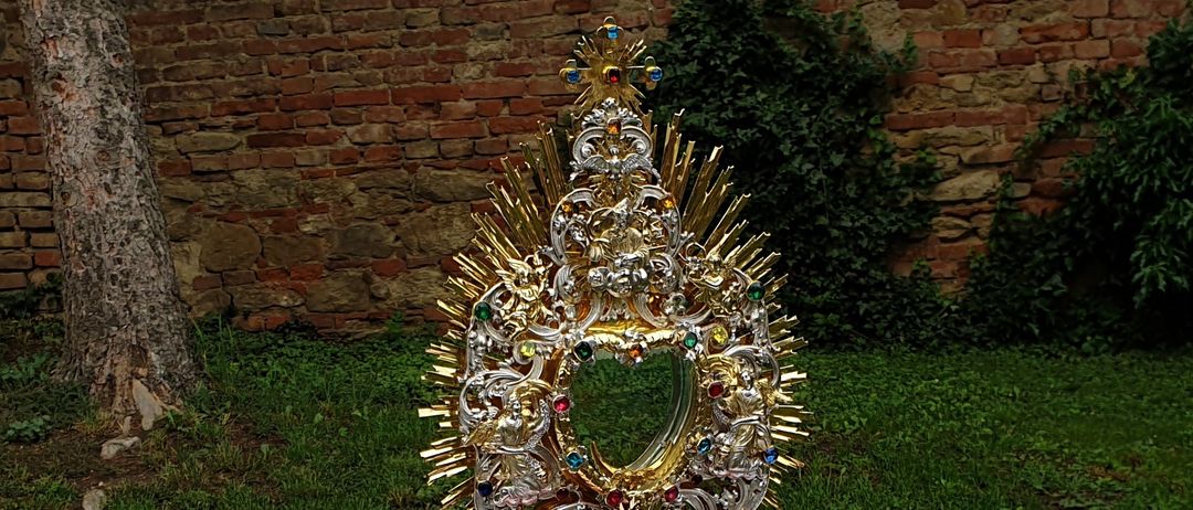 Ein verzierter goldener Monstranz steht auf einem mit bunten Blütenblättern bedeckten Tisch. Eine Ziegelmauer mit Grün im Hintergrund.