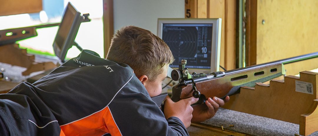 Ein junger Junge liegt auf dem Boden und zielt mit einem Gewehr auf ein Ziel, das auf einem Monitor angezeigt wird. Er trägt eine Jacke in Orange und Schwarz.