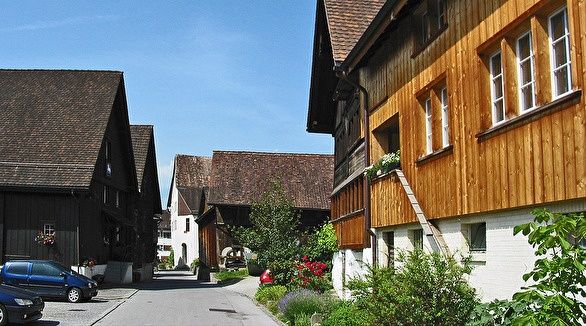 Eine ruhige Straße mit traditionellen Holzhäusern, einige mit Balkonen, unter einem klaren blauen Himmel. Ein blauer Van und ein rotes Auto sind geparkt.