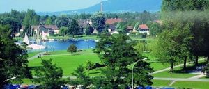 Eine Luftaufnahme einer Stadt mit einem See und angedockten Booten. Grüne Rasenflächen und Bäume umgeben den See. Eine Person fährt mit dem Fahrrad auf der Straße. Berge sind in der Ferne.