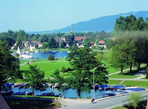 Eine Luftaufnahme einer Stadt mit einem See und angedockten Booten. Grüne Rasenflächen und Bäume umgeben den See. Eine Person fährt mit dem Fahrrad auf der Straße. Berge sind in der Ferne.