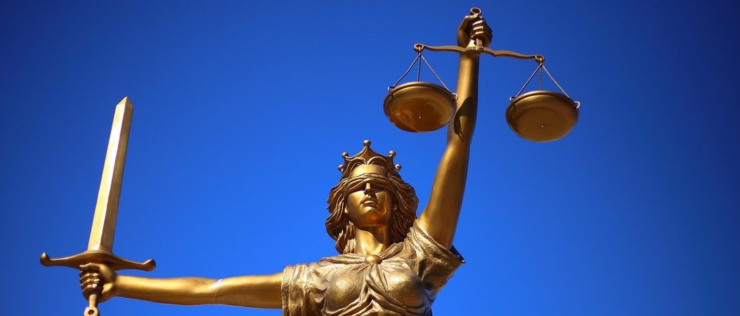 Eine goldene Statue einer verblindeten weiblichen Richterin, die eine Waage und ein Schwert unter einem blauen Himmel hält.