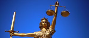 Eine goldene Statue einer verblindeten weiblichen Richterin, die eine Waage und ein Schwert unter einem blauen Himmel hält.
