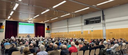Ein großer Saal im Innenraum beherbergt eine Versammlung mit einer Präsentation auf der Leinwand. Die Teilnehmer sitzen in Reihen und lauschen. Ein Podium mit Personen steht vorne.