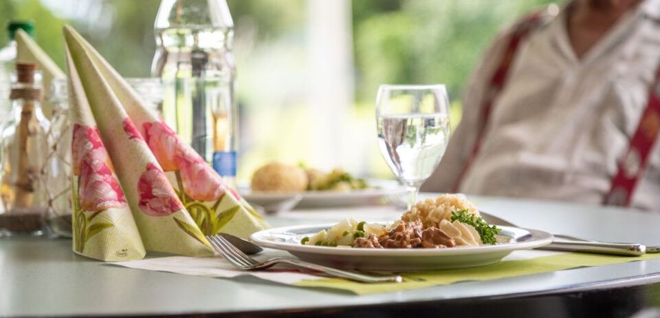 Ein gedeckter Tisch mit Essen, einem Glas Wasser und einer Flasche. Eine Person sitzt im Hintergrund.