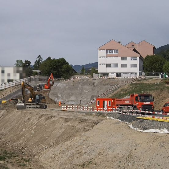 Eine Baustelle mit schwerem Gerät. Ein gelber Bagger und ein roter Lkw sind geparkt. In der Nähe steht ein Gebäude, und eine Person in einer orangefarbenen Weste geht vorbei.