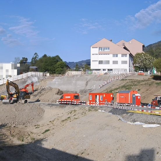 Eine Baustelle mit orangefarbenen Maschinen, einem weißen Gebäude und einem blauen Himmel mit Wolken. Im Hintergrund befinden sich Bäume und Berge.