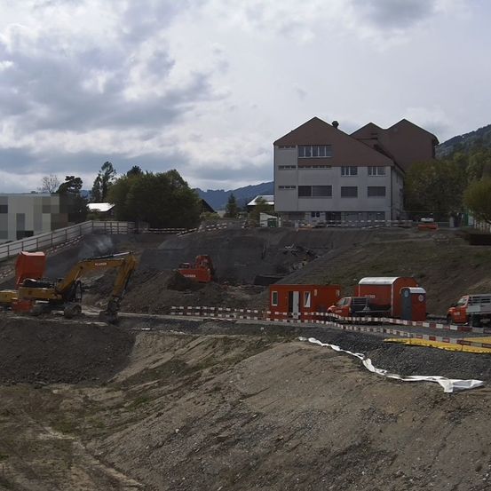 Ein Baugelände mit schweren Maschinen und Lastwagen unter einem bewölkten Himmel. Ein Bagger arbeitet an einem Hang mit einem großen Gebäude dahinter.